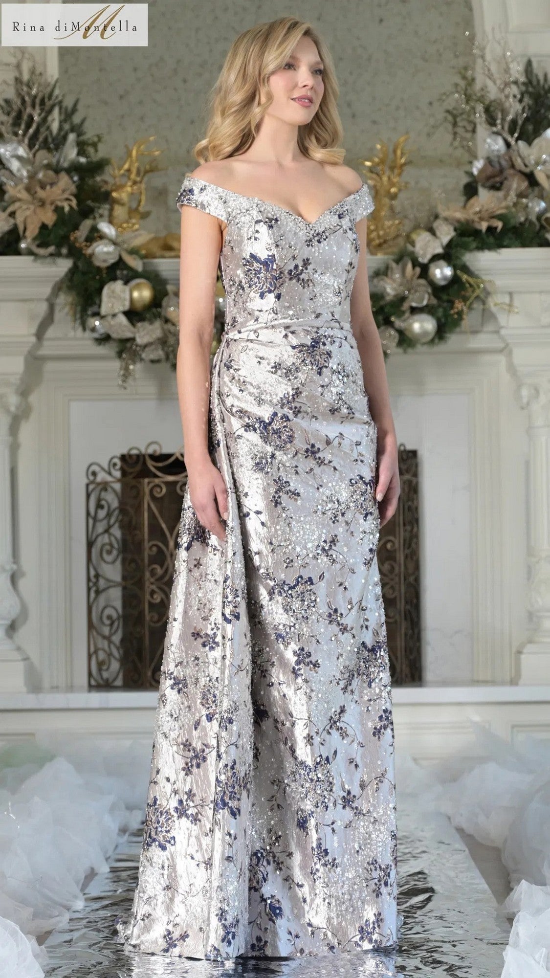 A woman is indoors wearing the Long Formal Dress: Rina Di Montella RD2912 in Navy Multi, featuring off-the-shoulder jacquard with silver and blue floral patterns. Holiday decorations and a white fireplace are in the background.